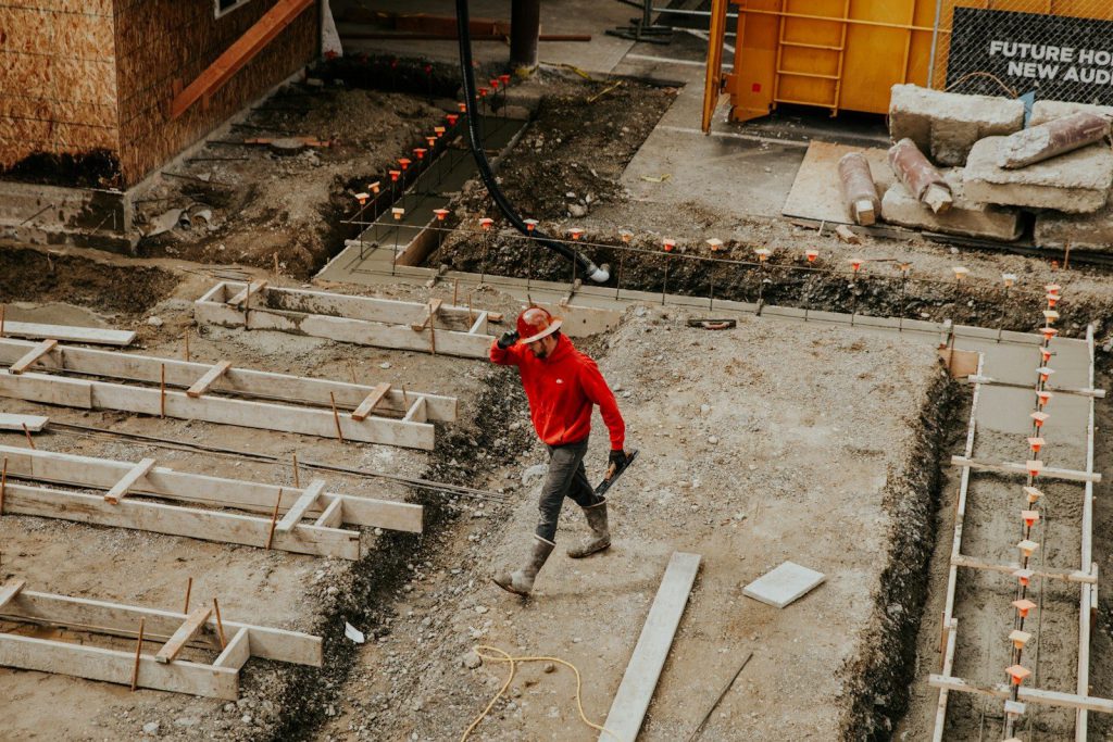 Công trường đang làm móng, các rãnh móng đã đào và lắp cốp pha gỗ, cốt thép chờ có chụp nhựa màu cam, một người thợ đội mũ bảo hộ đi giữa các rãnh